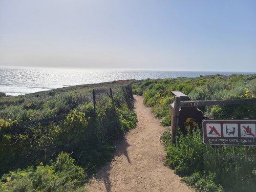 A sandy path leads to the ocean, surrounded by greenery and signs indicating area rules and hours.