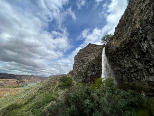 A waterfall cascades down a rocky cliff, surrounded by lush greenery and a cloudy sky in a scenic landscape.