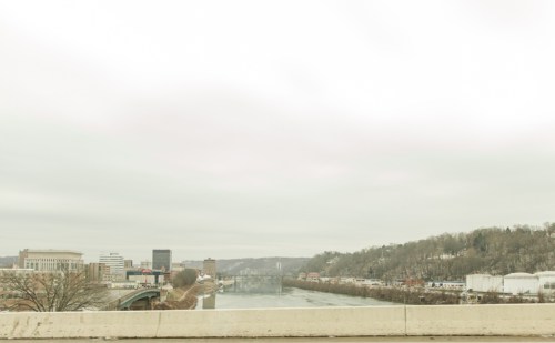 A cloudy view of a river with buildings and hills along the banks, seen from a bridge.