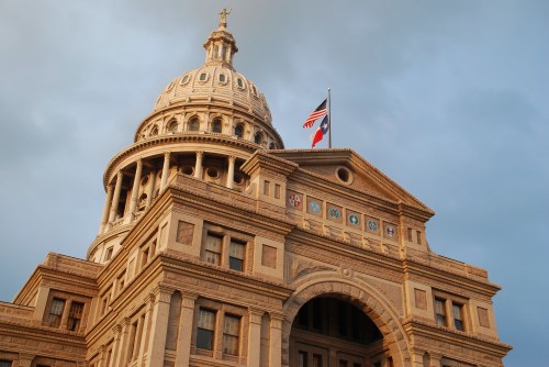 The Texas State Capitol building with a dome, featuring the American and Texas flags against a cloudy sky.