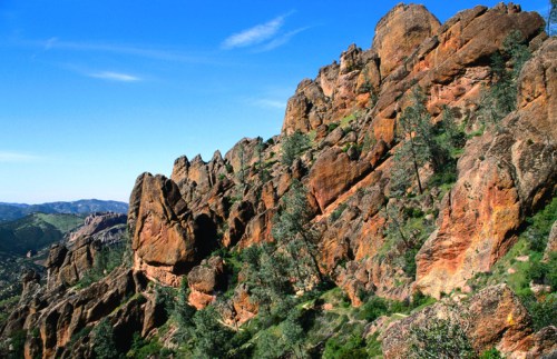 Rocky landscape with steep cliffs, scattered trees, and a clear blue sky in the background.