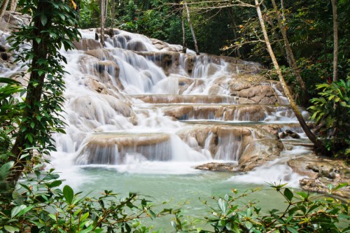 A serene waterfall cascades over rocky tiers, surrounded by lush greenery in a tropical forest.
