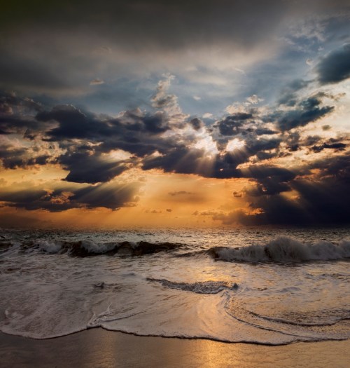 A dramatic sunset over the ocean, with rays of light breaking through dark clouds and waves gently lapping at the shore.