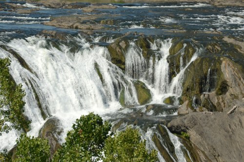 A cascading waterfall flows over rocky terrain, surrounded by greenery and a serene river landscape.