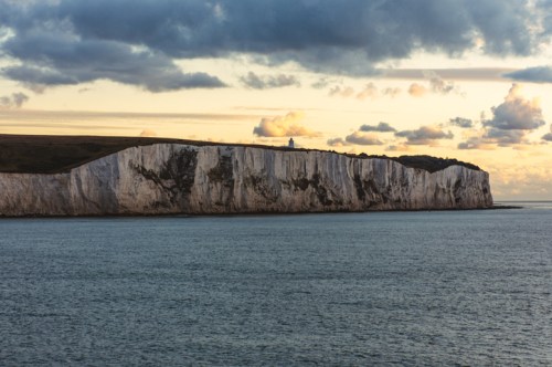 Cliffs rise above calm waters under a cloudy sky, with a lighthouse visible in the distance at sunset.