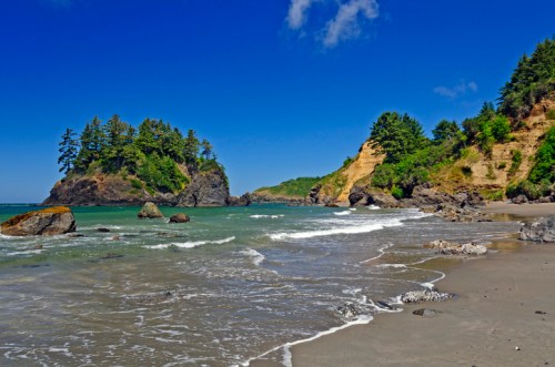 Scenic beach view with rocky cliffs, lush greenery, and gentle waves under a clear blue sky.