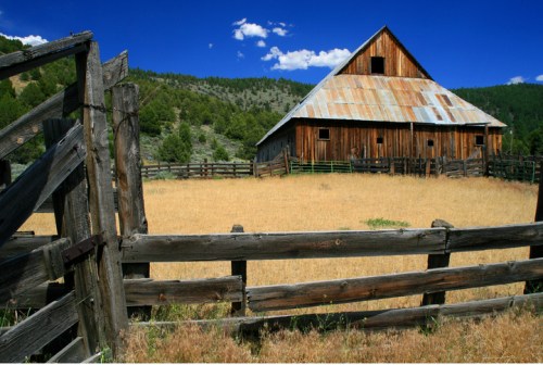 A rustic wooden barn with a metal roof, surrounded by a dry field and a wooden fence, set against a blue sky.