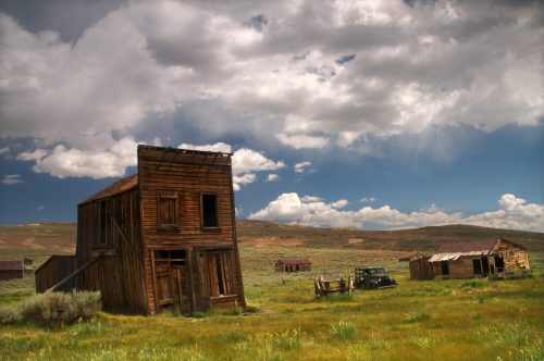 Abandoned wooden buildings in a grassy landscape under a cloudy sky, evoking a ghost town atmosphere.