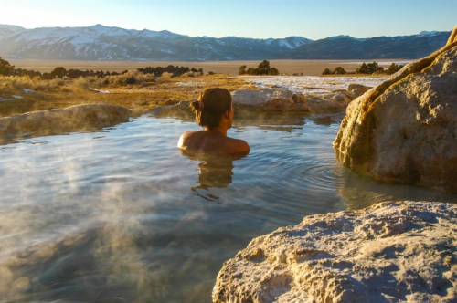 A person relaxes in a steaming hot spring, surrounded by rocks and mountains under a clear sky.