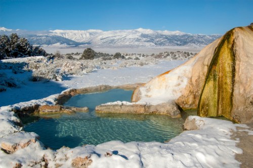 A serene hot spring surrounded by snow-covered mountains and a clear blue sky.