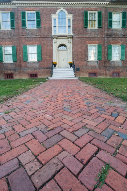 A brick pathway leads to a historic brick building with green shutters and a central door, flanked by two windows.