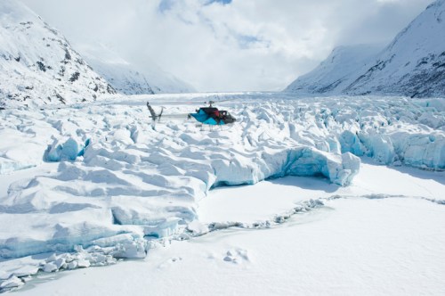 A helicopter rests on a snowy glacier, surrounded by icy formations and mountains under a cloudy sky.
