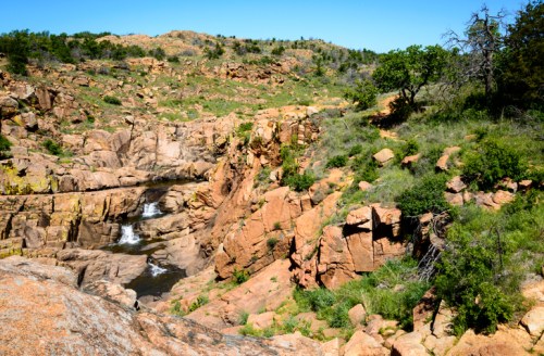 A rocky landscape with a small waterfall, lush greenery, and a clear blue sky in the background.