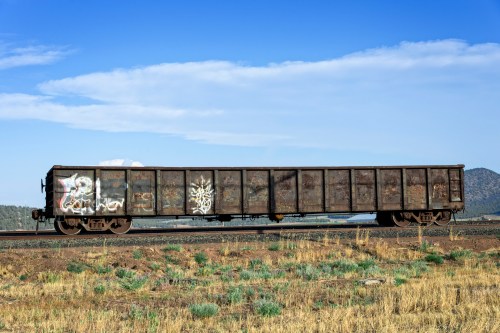 A long, empty freight train car with graffiti, set against a blue sky and distant mountains.