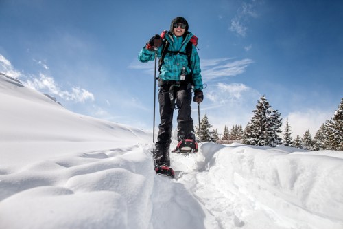 A person snowshoeing through a snowy landscape, wearing a blue jacket and smiling against a clear blue sky.