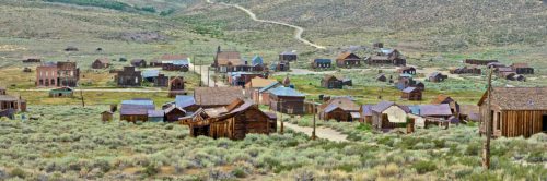 A panoramic view of a deserted ghost town with wooden buildings surrounded by sagebrush and rolling hills.