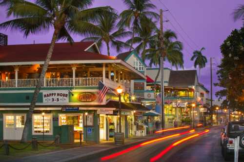 Colorful buildings line a street with palm trees, illuminated by evening lights and a purple sky at dusk.