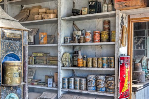 Shelves filled with vintage cans, jars, and containers in a rustic setting, showcasing old-fashioned goods and packaging.