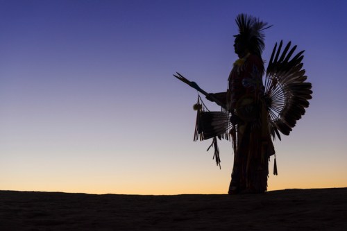 A silhouette of a person in traditional attire with feathers, holding a staff against a colorful sunset sky.