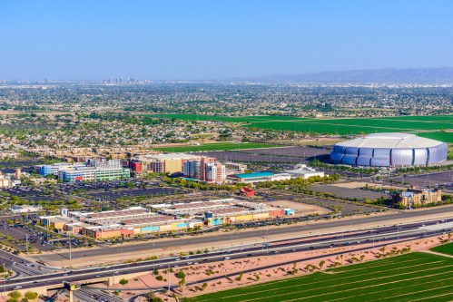 Aerial view of a cityscape with a stadium, commercial buildings, and green fields under a clear blue sky.