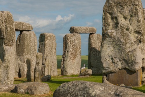 A view of Stonehenge with large stone pillars and a blue sky in the background. A person walks among the stones.