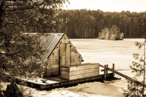 A rustic wooden cabin on a frozen lake, surrounded by trees and distant islands, captured in sepia tones.