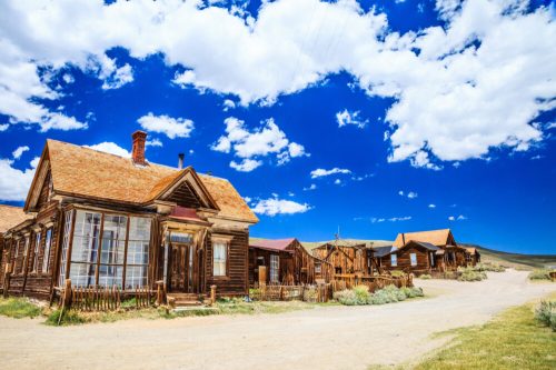 Abandoned wooden buildings under a bright blue sky with fluffy clouds, surrounded by sparse vegetation.