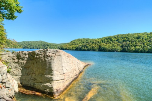 A serene lake surrounded by lush green hills and a large rock formation in the foreground under a clear blue sky.