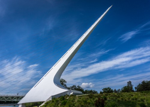 A tall, white, modern bridge structure with a sharp, angled design against a blue sky and green landscape.