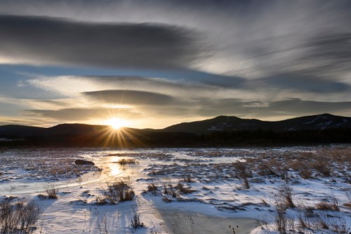 Sunset over a snowy landscape with mountains in the background and dramatic clouds in the sky.