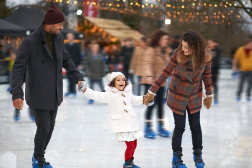 A family ice skating together, holding hands and smiling, with festive lights in the background.