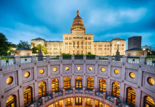 The Texas State Capitol building illuminated at dusk, viewed from a circular stone structure with arched windows.