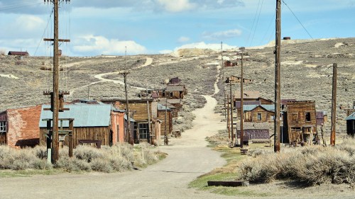 Abandoned ghost town with wooden buildings, dirt road, and power lines under a blue sky with scattered clouds.