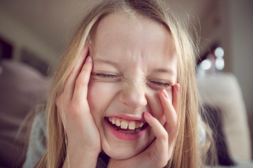 A joyful young girl with long hair smiles widely, covering her face with her hands in a playful expression.