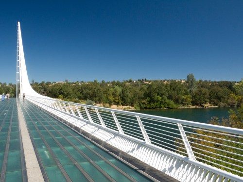 A modern white bridge spans over a river, surrounded by greenery under a clear blue sky.