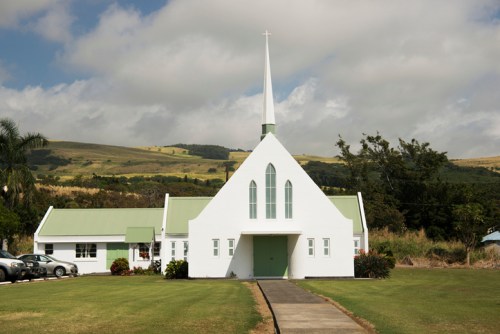 A white church with a green roof and tall spire, set against a backdrop of rolling hills and cloudy skies.
