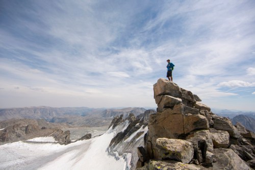 A person stands on a rocky peak, overlooking a vast mountainous landscape under a cloudy sky.