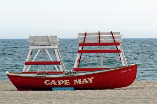 A red lifeguard boat labeled "Cape May" sits on a sandy beach with the ocean in the background.