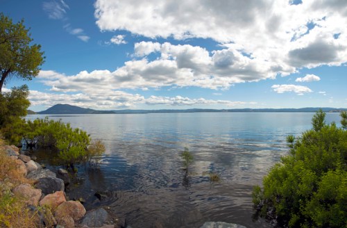 A serene lake scene with calm waters, lush greenery, and a distant mountain under a partly cloudy sky.