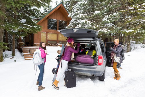 Three friends unpacking a car in the snow, with a cozy cabin in the background and winter gear scattered around.