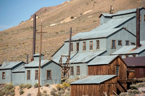 Abandoned mining buildings with metal roofs, set against a rugged, hilly landscape.