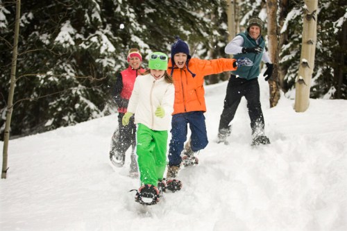 A group of four children joyfully snowshoeing through a snowy forest, wearing colorful winter clothing.