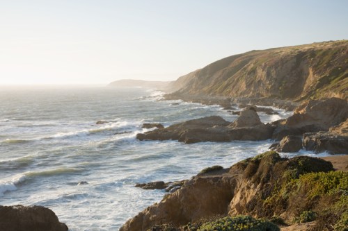 A scenic coastal view with waves crashing against rocky cliffs under a clear sky.