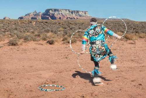 A dancer in traditional attire performs with hoops against a desert landscape and distant rock formations.