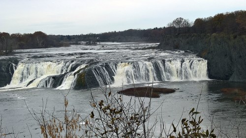 A scenic view of a waterfall cascading over rocky cliffs into a calm river, surrounded by trees and autumn foliage.