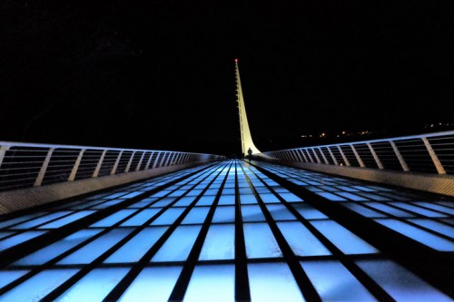 A lit bridge at night, featuring a glowing blue pathway and a tall, illuminated spire in the background.