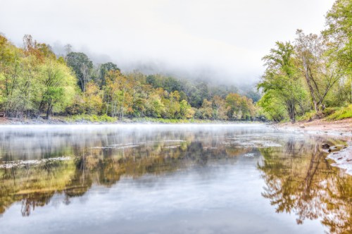 A serene river scene with mist, surrounded by trees in autumn colors and a calm, reflective surface.
