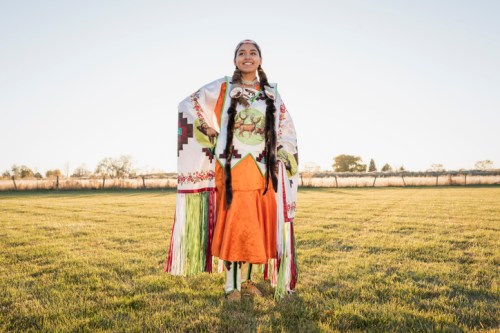A young Indigenous woman stands proudly in traditional attire on a grassy field, with a bright sky in the background.