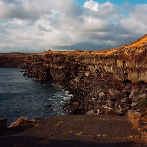 Rocky coastline under a cloudy sky, with cliffs rising above the calm sea and patches of dry grass along the shore.