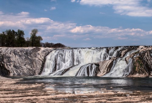 A scenic waterfall cascading over rocky cliffs, surrounded by trees and a blue sky with fluffy clouds.
